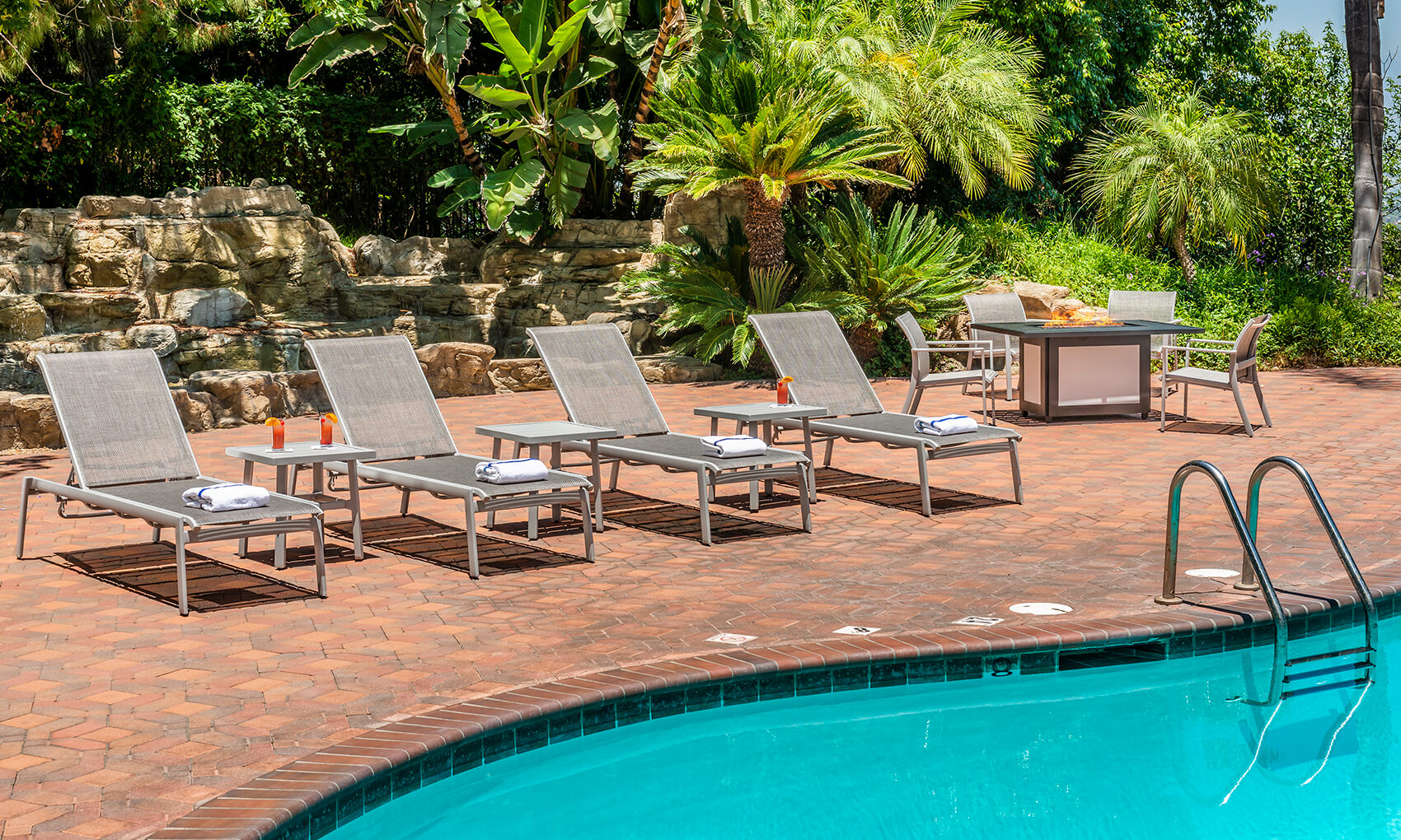 Four Gensun Echelon lounge chairs by a swimming pool, with towels and a scenic, lush garden background. A small round table and additional patio seating are visible near the pool.