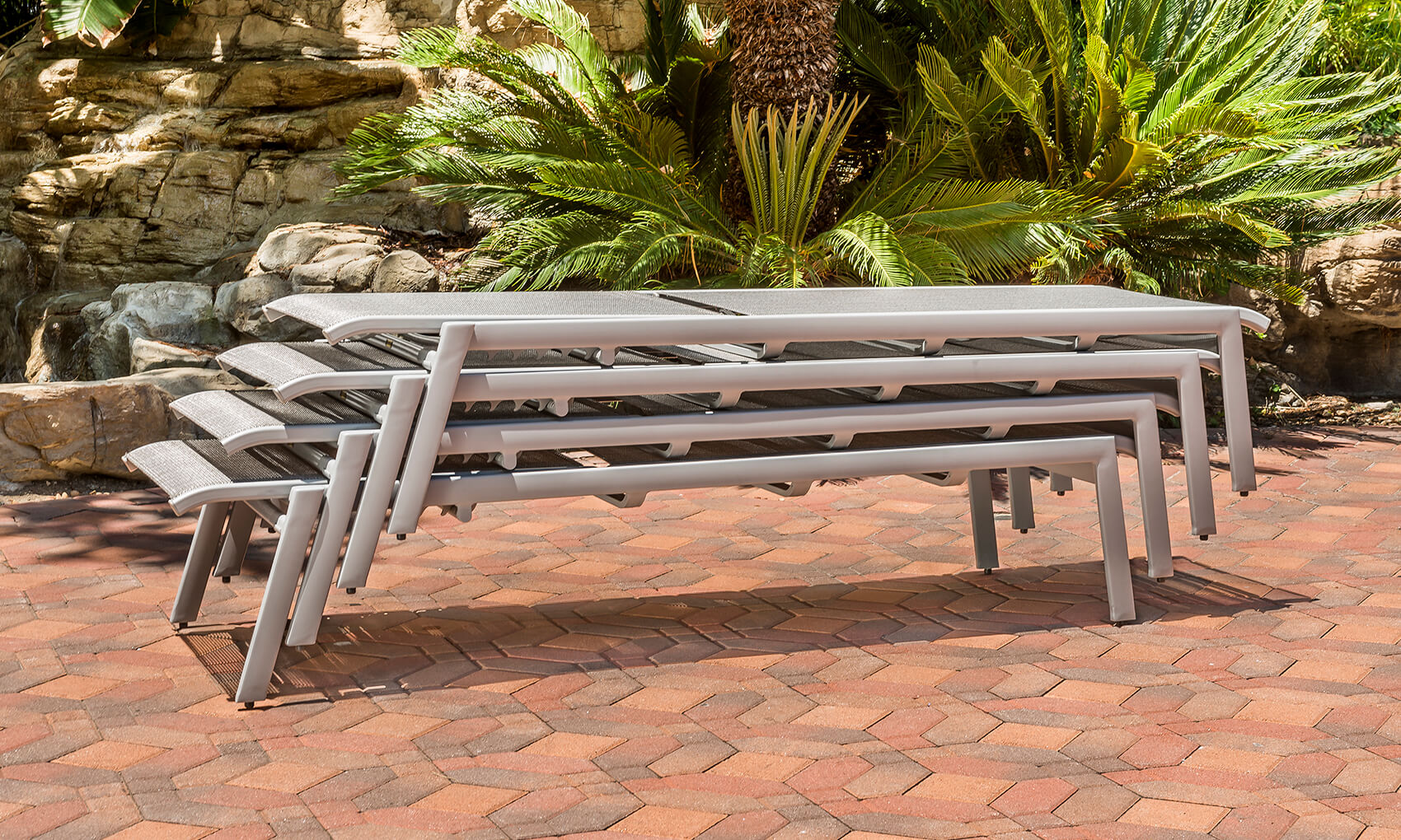 Four gray Gensun Echelon metal benches stacked in a neat row on a red brick patio, surrounded by lush greenery and a rocky landscaped backdrop in a park setting.