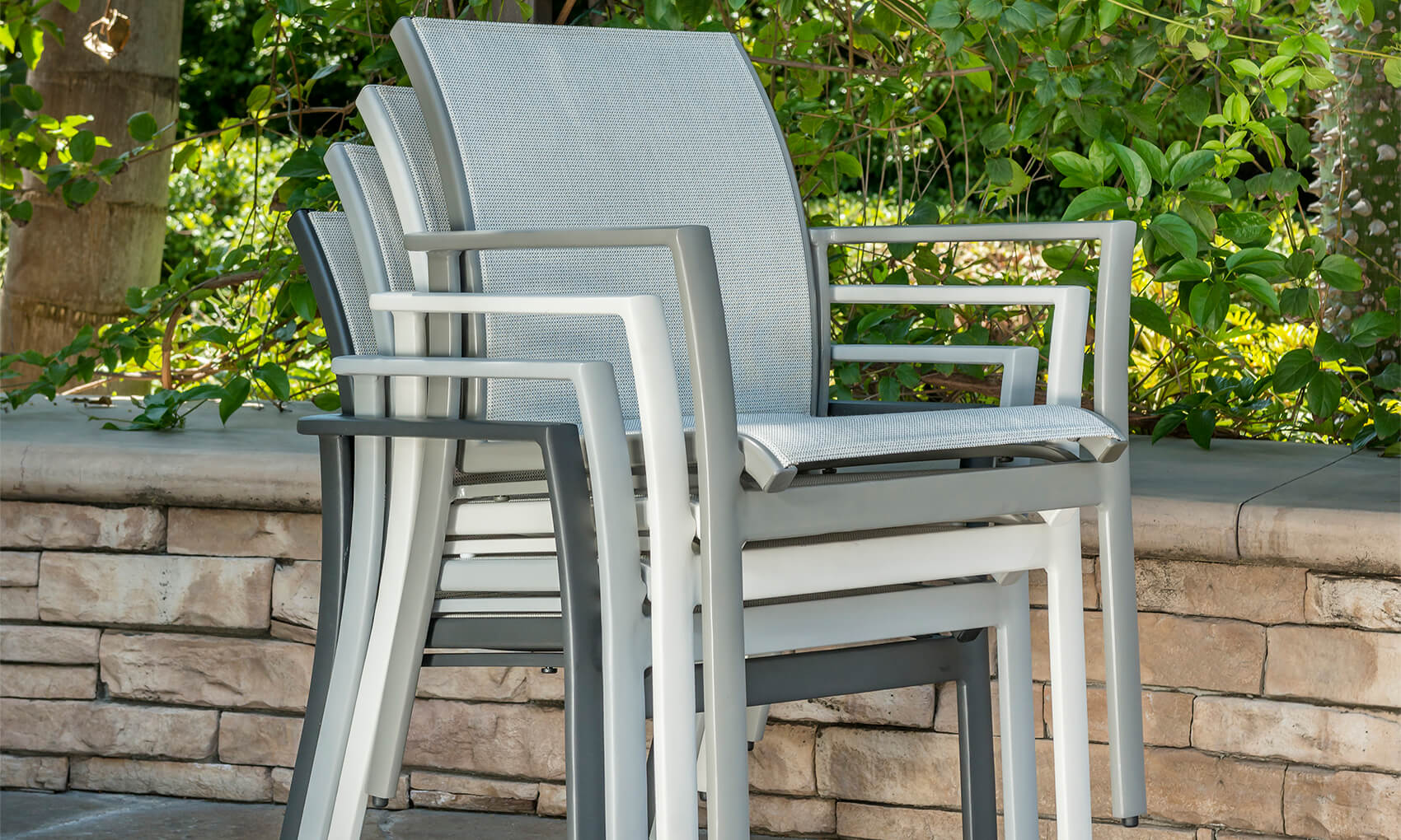 A stack of Gensun Echelon metal patio chairs with gray woven backrests and seats, placed beside a low stone wall surrounded by green foliage in an outdoor garden setting.