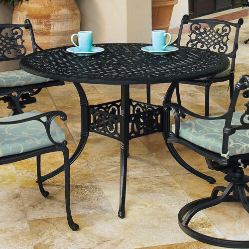 An outdoor seating area with a round black Gensun Michigan wrought iron table and four matching chairs, each with a cushioned seat. Two teal cups are placed on the table.
