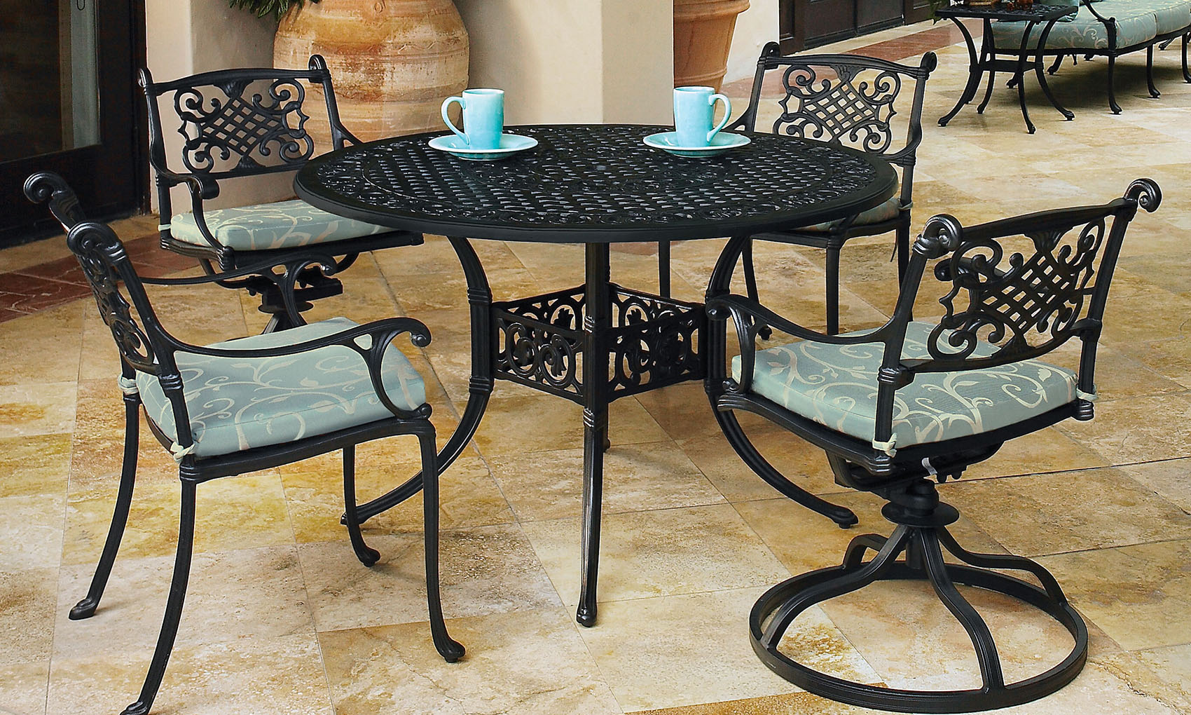 An outdoor seating area with a round black Gensun Michigan wrought iron table and four matching chairs, each with a cushioned seat. Two teal cups are placed on the table.