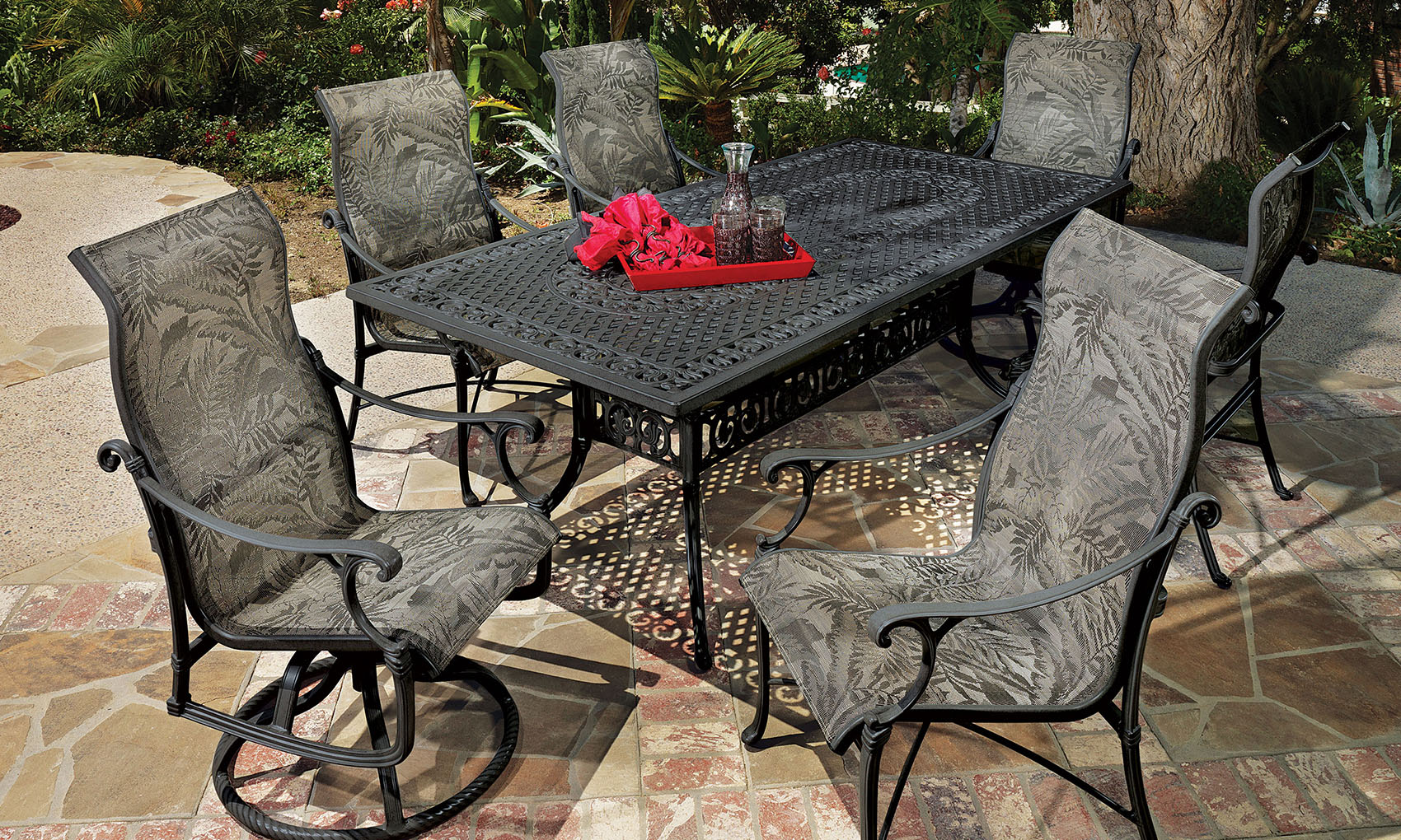 A Gensun Michigan patio dining set featuring a large rectangular metal table and six cushioned chairs in a garden, with a tray on the table under a sunny ambiance.