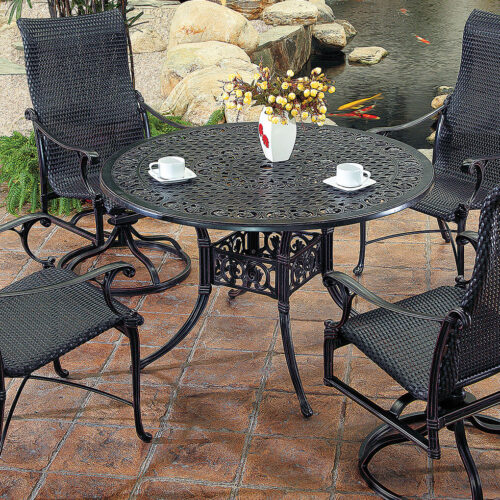 An outdoor Gensun Michigan patio set with four black, metal, mesh chairs surrounding a matching circular table, displayed on a stony surface. The table hosts a floral centerpiece and two white cups.