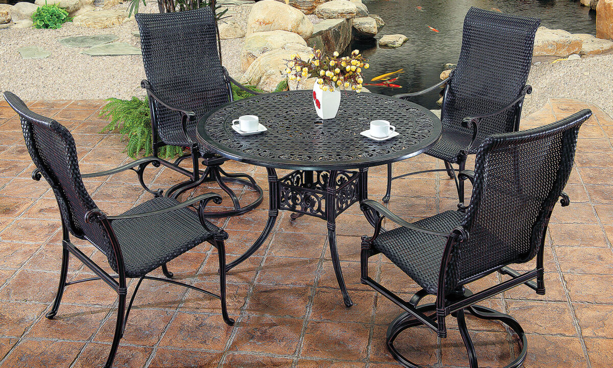 An outdoor Gensun Michigan patio set with four black, metal, mesh chairs surrounding a matching circular table, displayed on a stony surface. The table hosts a floral centerpiece and two white cups.