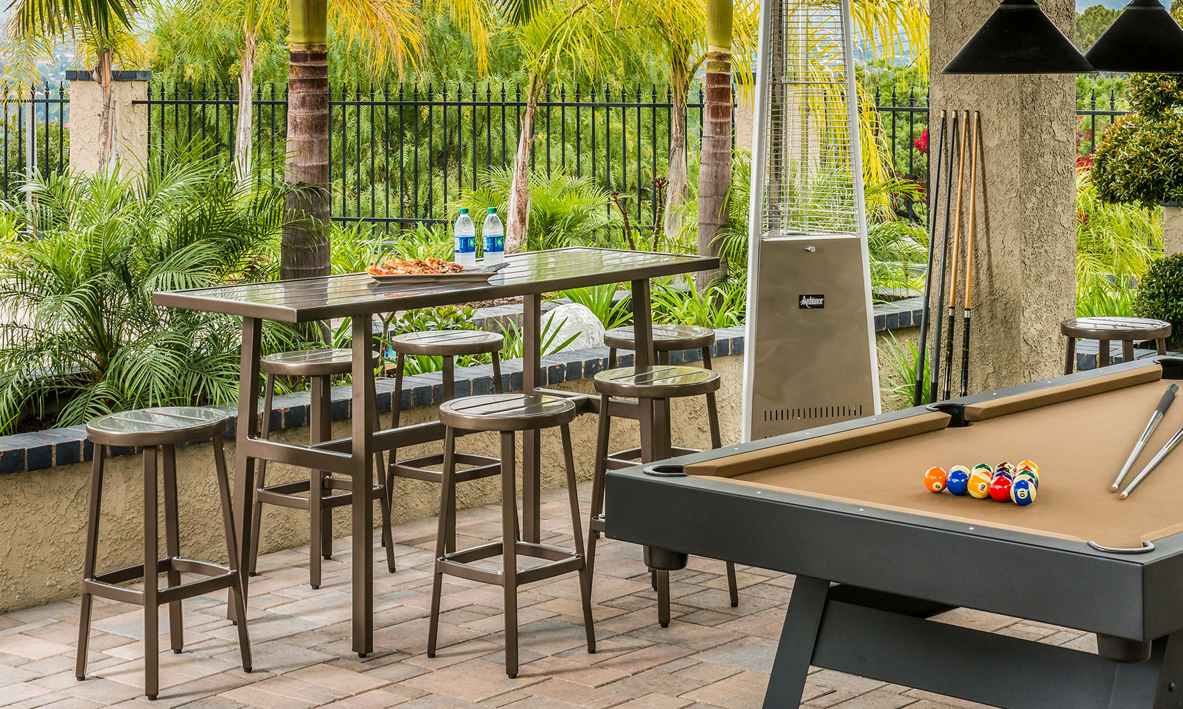 An outdoor entertainment space featuring a Gensun Plank pool table with balls set for a game, a tall bar table with stools, and a gas grill, all set against a backdrop of lush palms