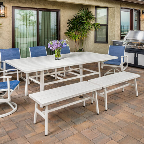An outdoor patio with a Gensun Plank dining set consisting of a white table and blue chairs, surrounded by potted plants and an equipped grill station.