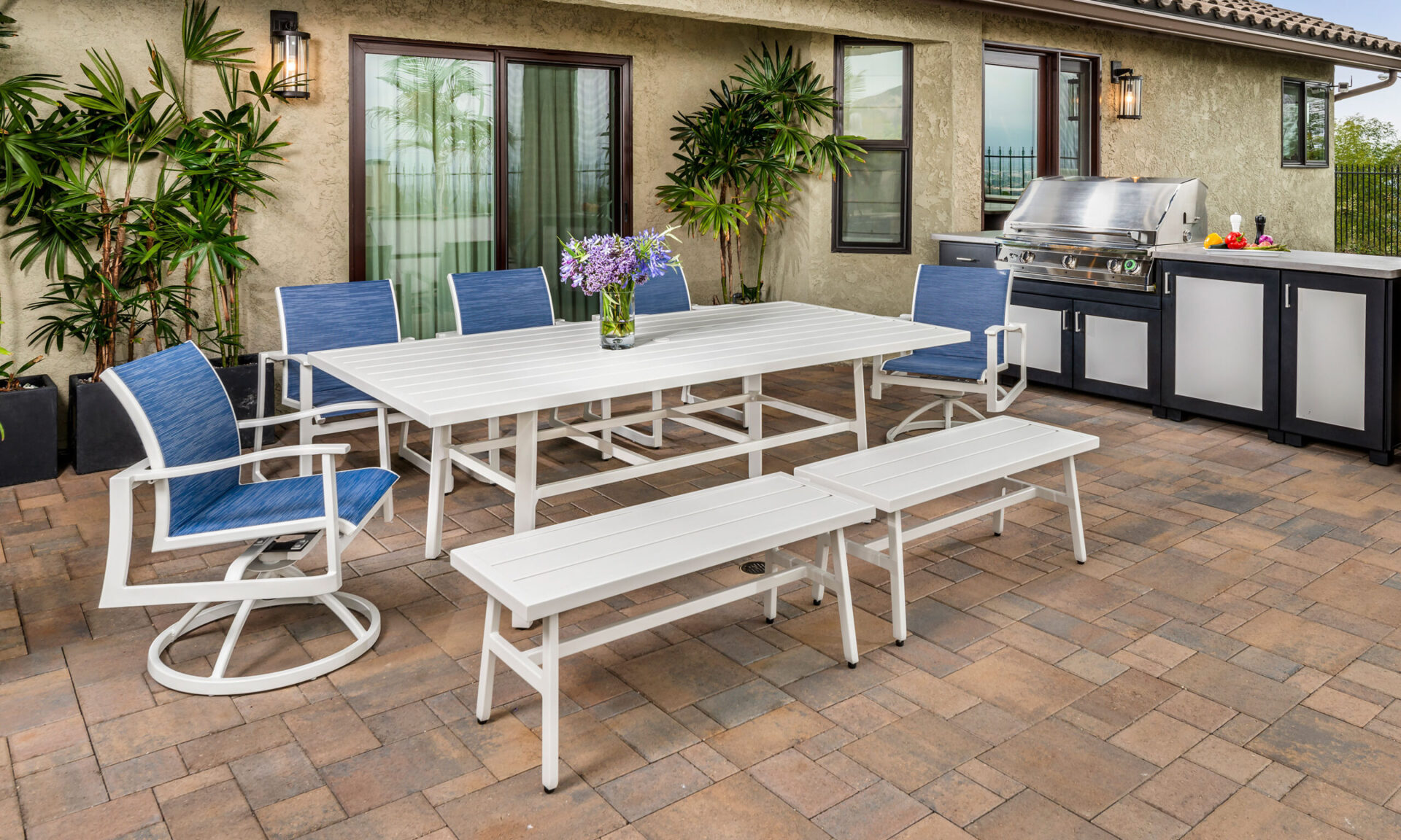 An outdoor patio with a Gensun Plank dining set consisting of a white table and blue chairs, surrounded by potted plants and an equipped grill station.