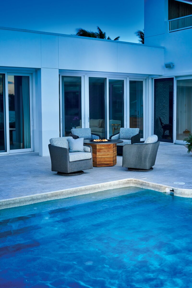An inviting outdoor seating area next to a swimming pool at dusk, featuring a fire pit center table, surrounded by four plush armchairs, adjacent to a modern white building.