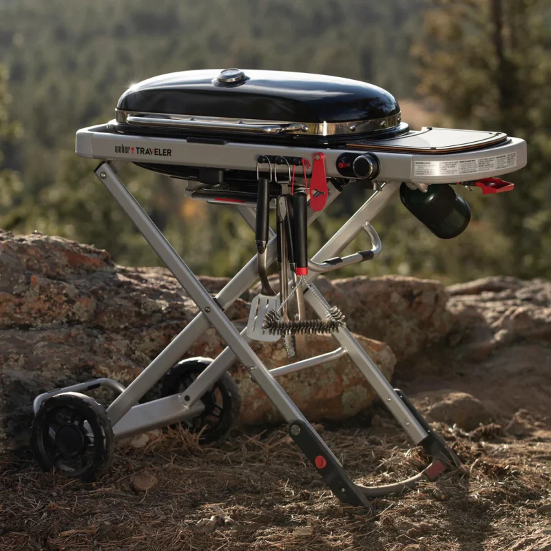 A portable Weber Traveler grill stands on a foldable cart in a wilderness setting, surrounded by woods and rocks under a clear sky.