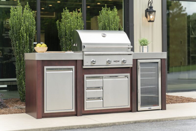 Outdoor kitchen island featuring a stainless steel grill station with a refrigerator and storage drawers built into a wooden base, displayed in front of a glass building surrounded by greenery.