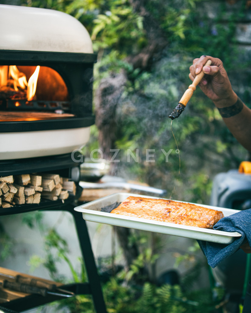 A person using a peel to slide a freshly baked focaccia bread into a wood-fired oven in a lush garden setting, with visible flames inside the oven and chopped wood stacked nearby.