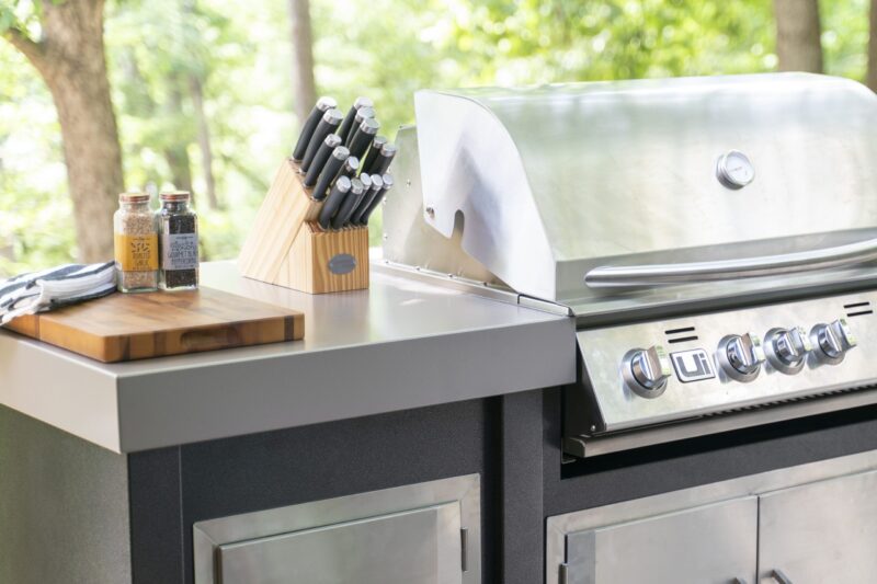 An outdoor kitchen island featuring a shiny stainless steel gas grill, a block of kitchen knives, and two jars of spices on a wooden countertop surrounded by green trees.