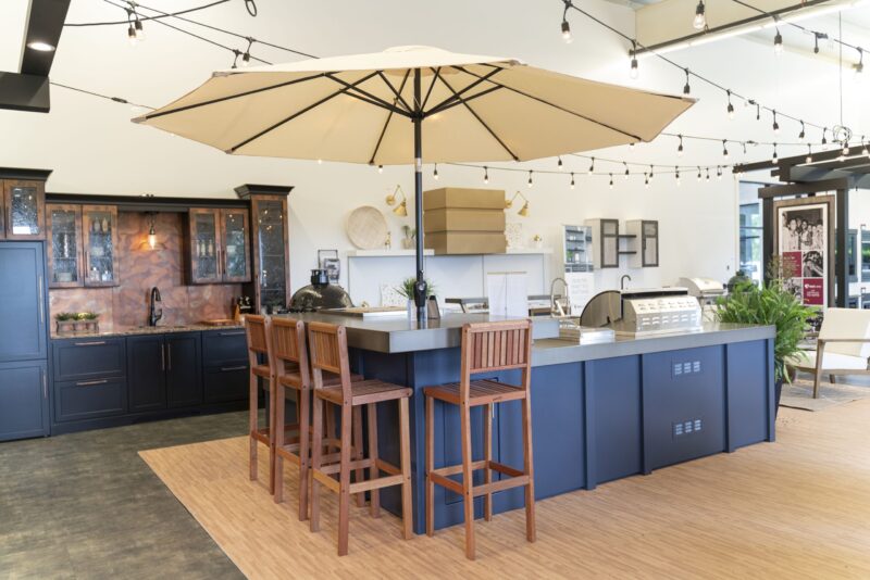 A modern and spacious outdoor kitchen showroom featuring dark blue cabinetry, wooden stools at a central island, overhead lighting, and a large beige umbrella. Bright and neatly organized, with various kitchen appliances visible.