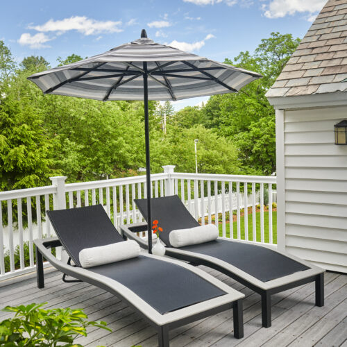 Two Bazza lounge chairs with cushions under an open umbrella on a wooden deck near green trees and a white house, suggesting a tranquil outdoor relaxation area.