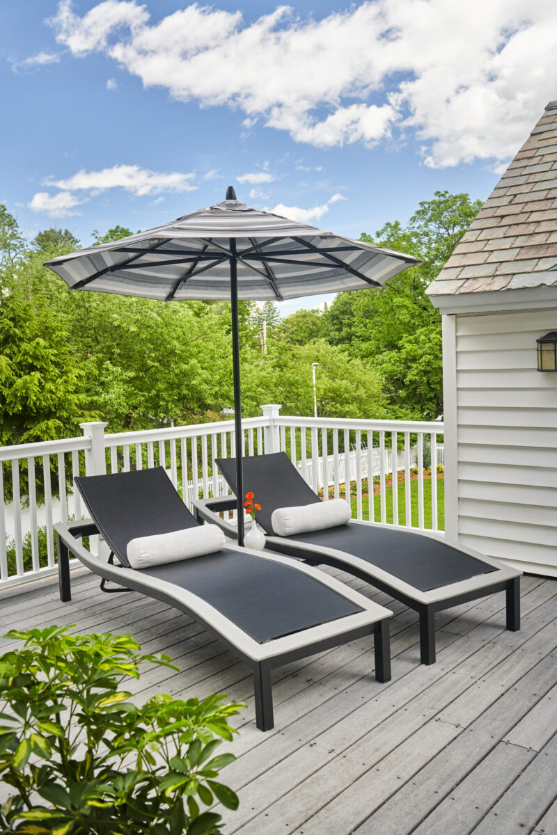 Two Bazza lounge chairs with cushions under an open umbrella on a wooden deck near green trees and a white house, suggesting a tranquil outdoor relaxation area.