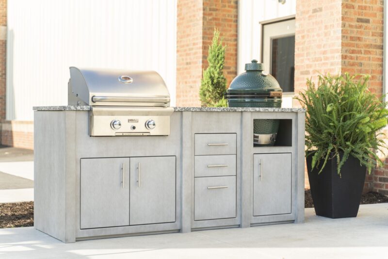 Outdoor kitchen island setup featuring a stainless steel grill and a ceramic smoker built into a concrete countertop with storage cabinets, next to a brick building and a potted plant.
