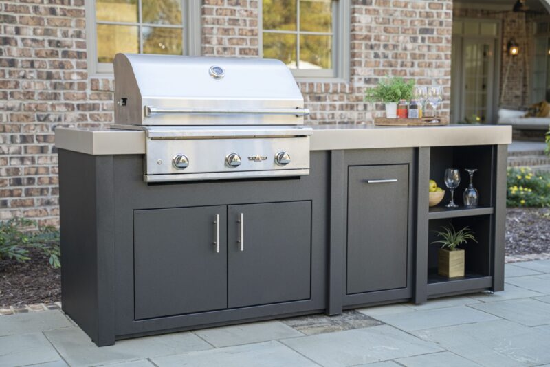 A modern outdoor kitchen island setup featuring a large stainless steel grill with storage compartments, set against a brick house with a visible patio area.