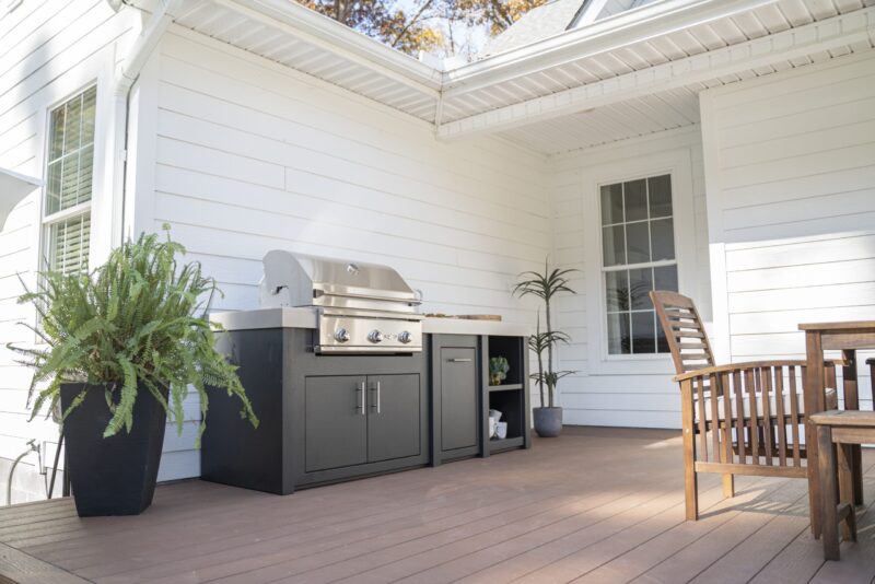 A spacious wooden deck features a modern outdoor kitchen island setup, including a large grill and cabinets. There's a wooden dining chair, with decor such as potted plants enhancing the cozy ambiance.