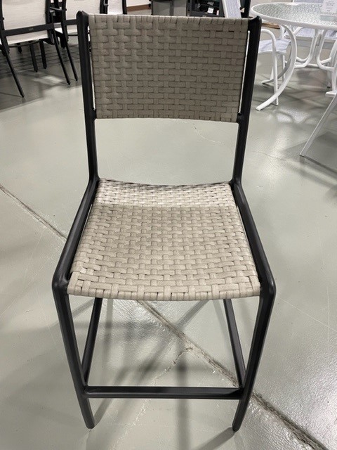 A modern Gensun Amari chair with a black metal frame and a light beige woven seat and backrest, standing on a polished concrete floor in a showroom setting.