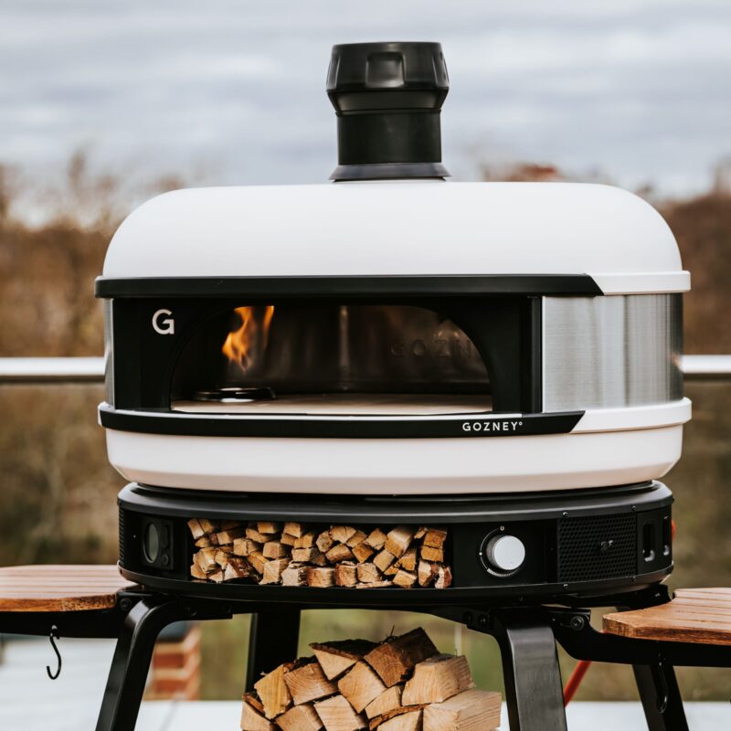An outdoor pizza oven with a sleek, modern design, featuring a visible fire and stocked wood compartment, set on a wooden table against a blurred natural backdrop.