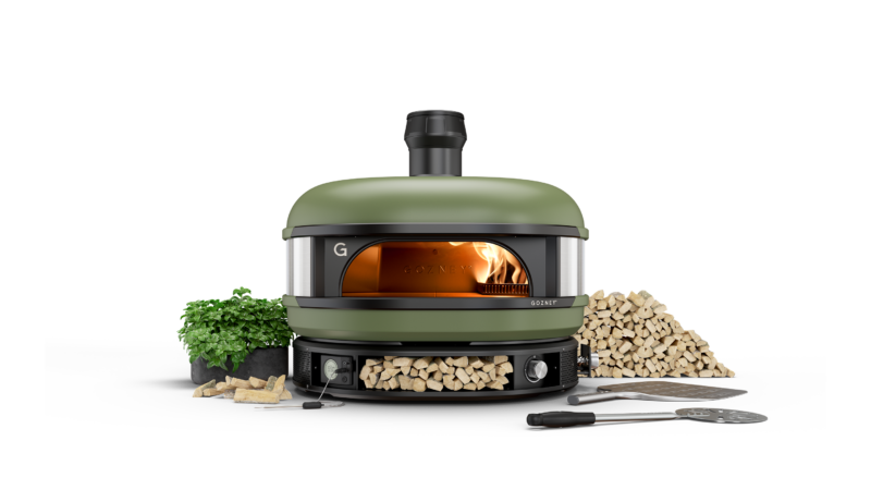 A modern green dome pizza oven with a visible flame inside, surrounded by firewood, a basil plant, and pizza-making tools on a black background.