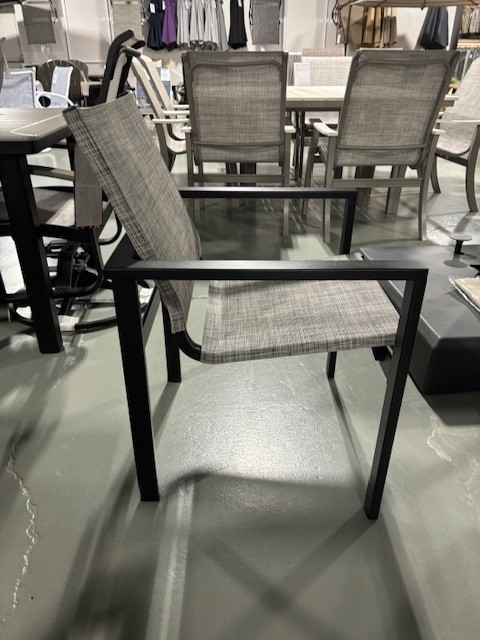 A modern Tribeca chair with a black metal frame and light gray woven fabric seat and backrest, displayed in a showroom with various furniture pieces in the background.