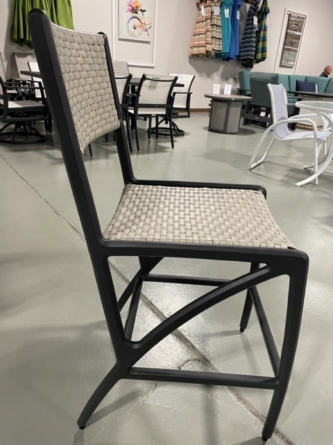 A modern Gensun Amari black chair with a woven beige seat and backrest, displayed in a store with various furniture in the background. The chair has a sleek, angular metal frame.