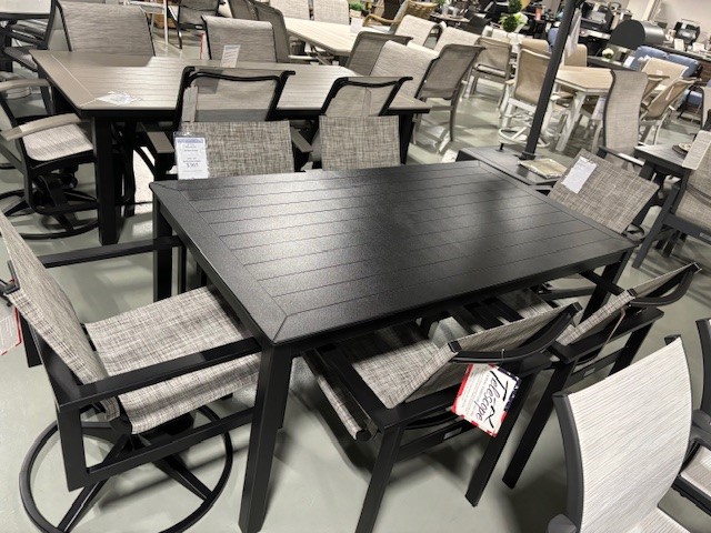 A display of Tribeca outdoor furniture featuring dark-framed tables and chairs with gray woven seat and backrest, set up in a store showroom with other furniture items in the background.