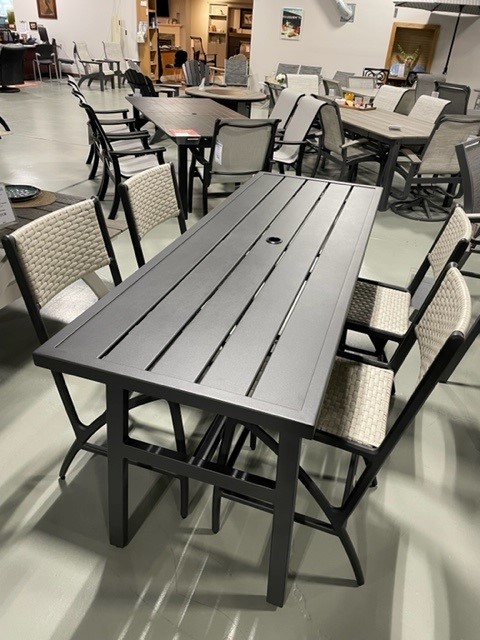 A showroom displaying a modern Gensun Amari outdoor dining set with a rectangular grey slatted table and surrounding wicker chairs with gray cushions, set on a concrete floor.