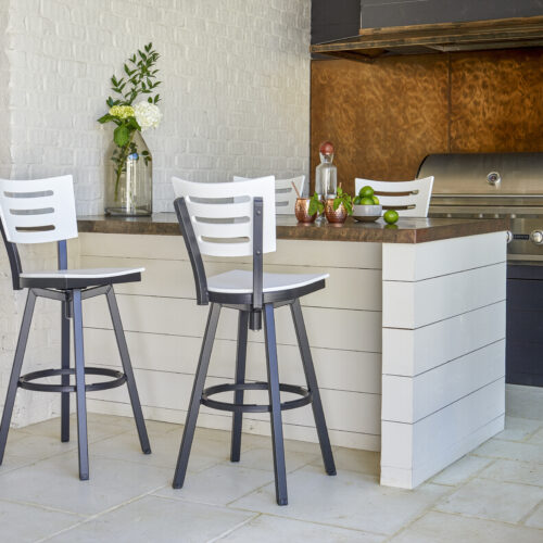 An avant-garde outdoor kitchen with a white brick wall, featuring a breakfast bar with two white and black bar stools and a copper backsplash. There are flowers and culinary herbs on the counter.