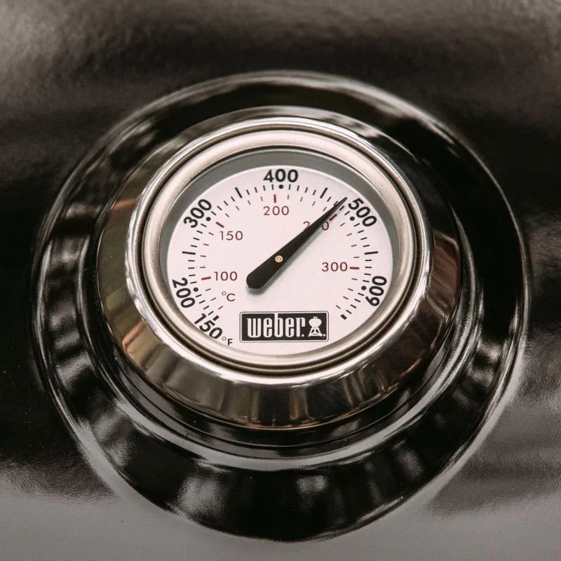 Close-up of a Weber Traveler grill thermometer embedded in a black lid, showing a temperature scale in both Fahrenheit and Celsius. The needle points slightly above 100 degrees Fahrenheit.