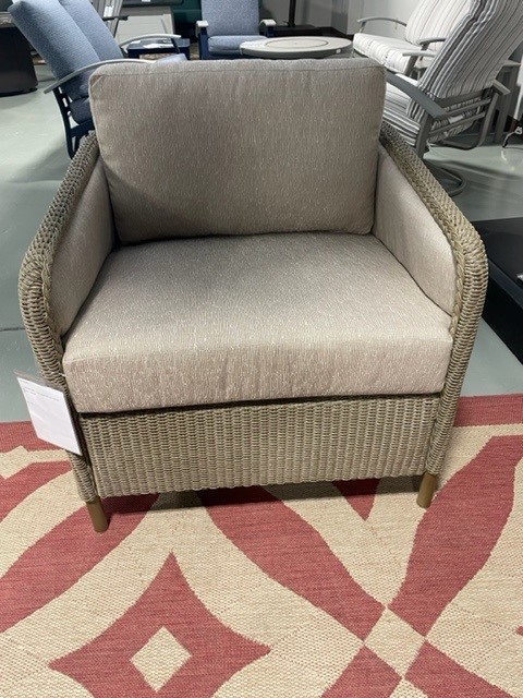 A comfortable, modern armchair with beige upholstery and a woven, natural-tone frame, displayed on a patterned area rug in a showroom setting.