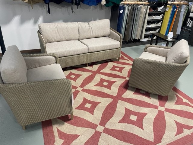 A modern living room setup featuring a beige sectional sofa and two matching armchairs on a red and beige patterned rug, displayed in a showroom with various fabrics in the background.