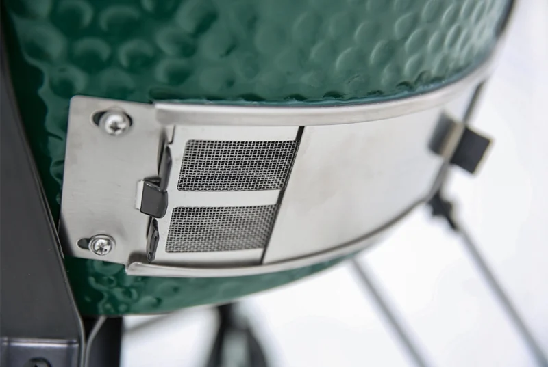 Close-up image of a Large Big Green Egg ceramic grill with a metal vent at the top, showcasing details of the vent's metal mesh and hinge mechanism against a blurred background.