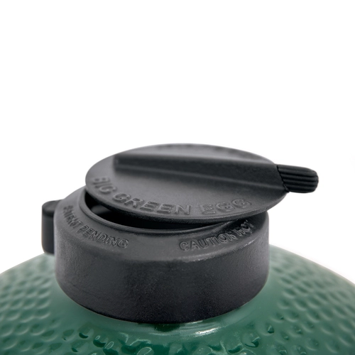 Close-up view of the top vent of a large Big Green Egg grill, showing the logo and caution text, set against a soft-focused background of the grill's textured green surface.