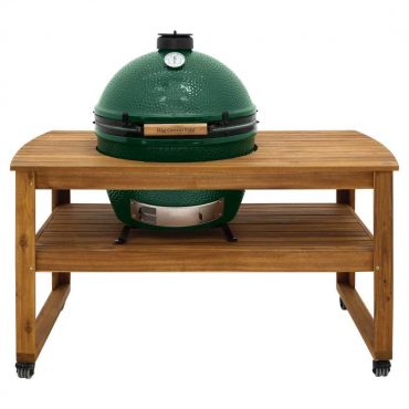 A Big Green Egg grill is perched on a wooden table with a lower shelf and wheels. It features a temperature gauge mounted on its green ceramic lid.