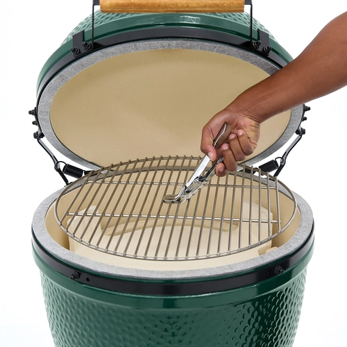 A person's hand using tongs to position a grill grate onto a Large Big Green Egg Package, featuring a ceramic, green-colored barbecue smoker. The smoker lid is open, revealing a clean, unused