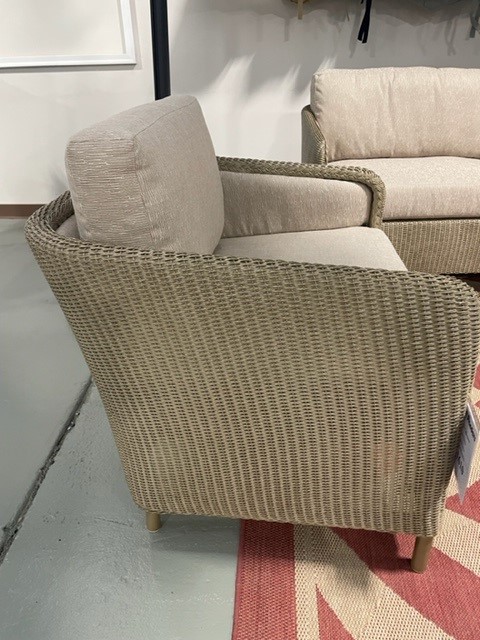 A beige woven wicker armchair with a cushioned seat and backrest, situated on a multicolored rug in an indoor setting.