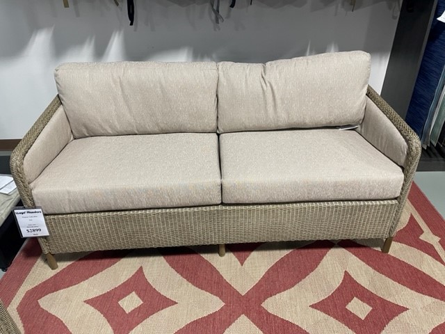 A beige two-seater sofa with woven side and back panels on a red and beige patterned rug, displayed in a showroom with a price tag visible.