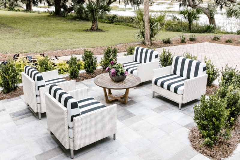 Outdoor patio area with custom patio sets including four white and navy striped armchairs around a round wooden table, surrounded by lush greenery and trees, set on a patterned tile floor.