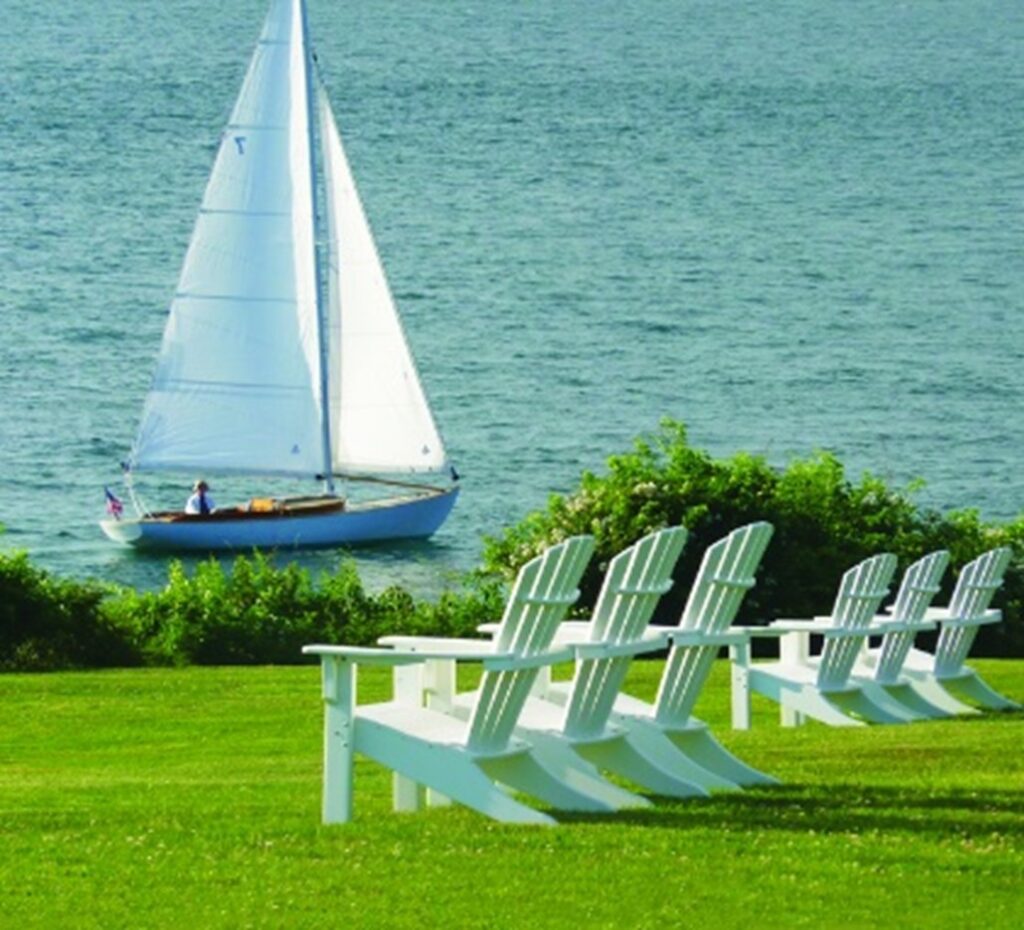 white shellback Adirondack chairs on green grass with a sailboat and water in the background