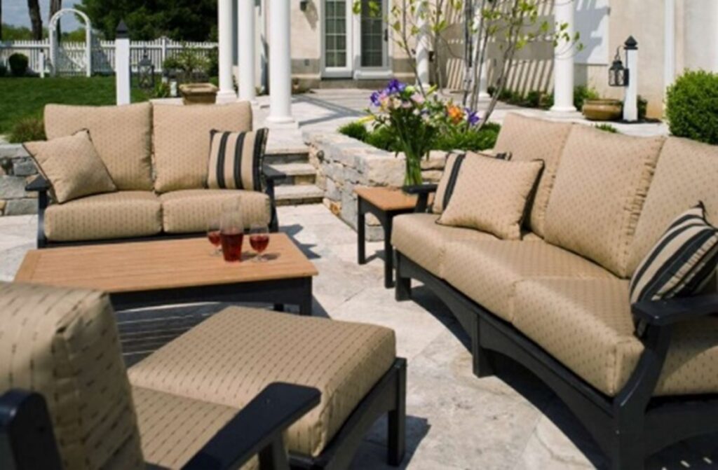 Breezesta deep seating sofa and loveseat with a coffee table and ottoman, tan cushions and black frames, stone flooring, floral planter in the background