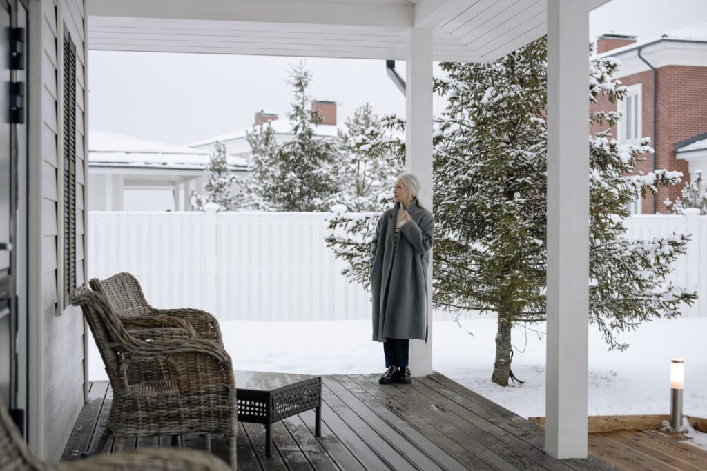 Woman in a gray coat standing on a porch with wicker furniture; snow on trees and the ground