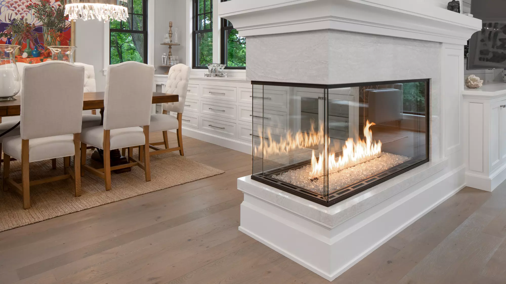 A modern, well-lit dining room with a large table and upholstered chairs features the Enlight Pier Three-Sided Gas Fireplace by Stellar by Heat & Glo prominently in the foreground, adding warmth to the