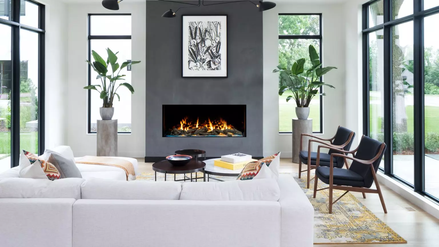 A modern living room featuring a white sofa, wooden chairs, and an Enlight Single-Sided Gas Fireplace by Stellar by Heat & Glo. Large windows provide ample natural light, with decorative plants and an abstract