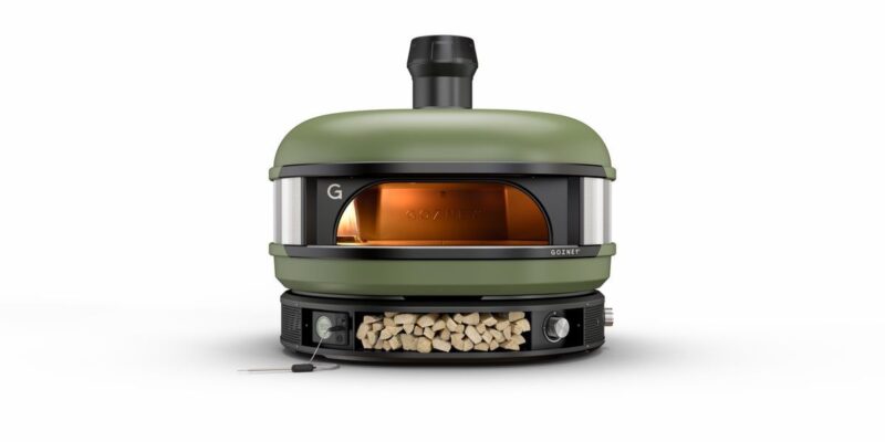 A modern, olive green outdoor pizza oven with a visible fire inside, featuring a front glass door and a chimney on top, set against a plain white background.