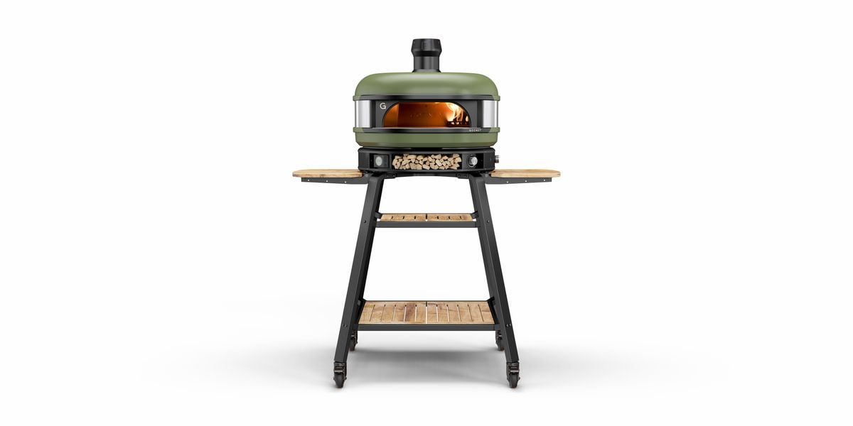 A modern green pizza oven with a visible fire chamber and wooden side shelves placed on an elevated metallic stand, against a plain white background.