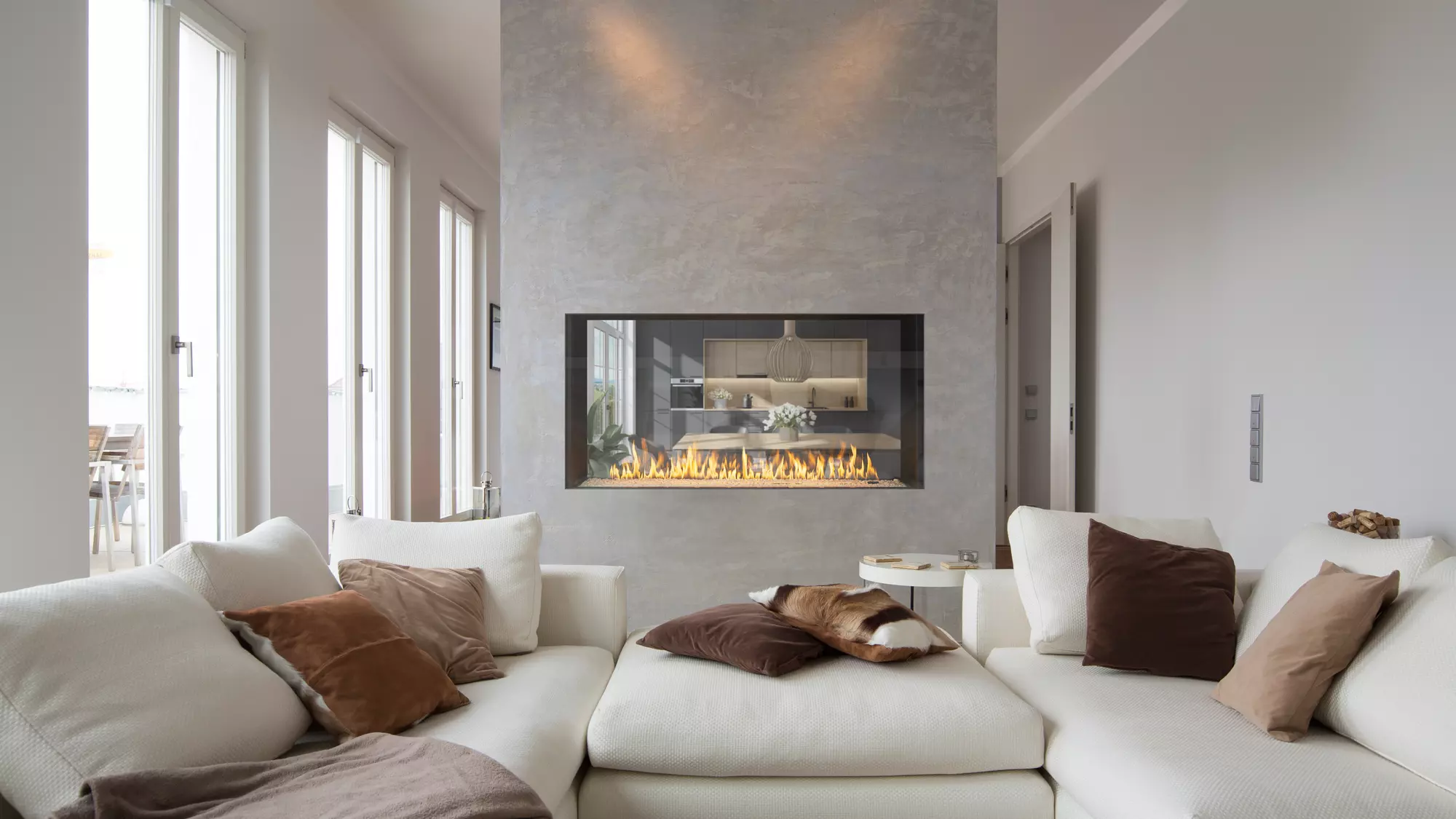 A cozy living room featuring a large white sectional sofa with brown and beige pillows, an Enlight See-Through Gas Fireplace by Stellar by Heat & Glo set in a concrete wall, and soft lighting. There