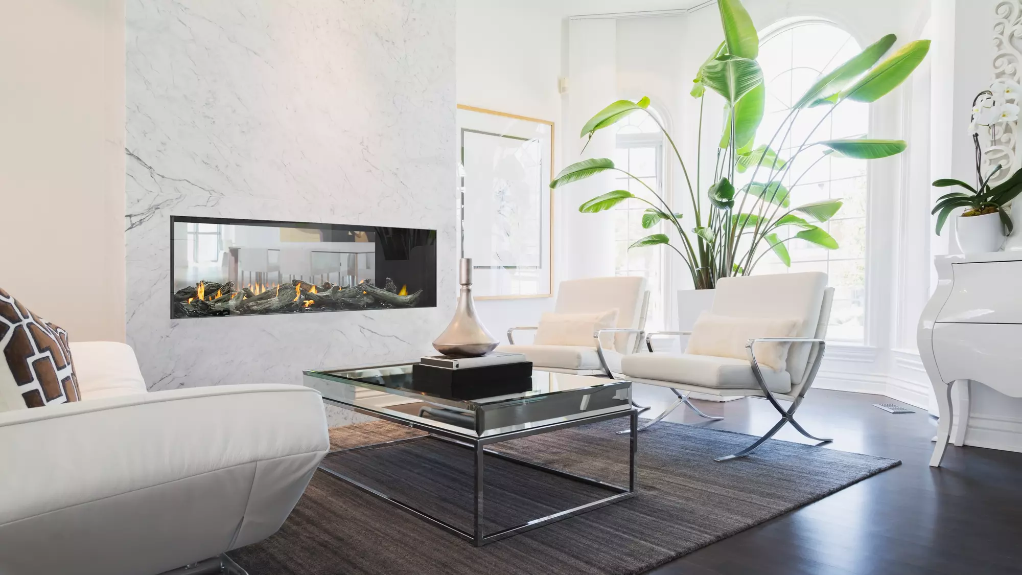 A sleek modern living room with white furniture and accents, an Enlight See-Through Gas Fireplace by Stellar by Heat & Glo, towering green plants, and a glass coffee table above a dark rug.