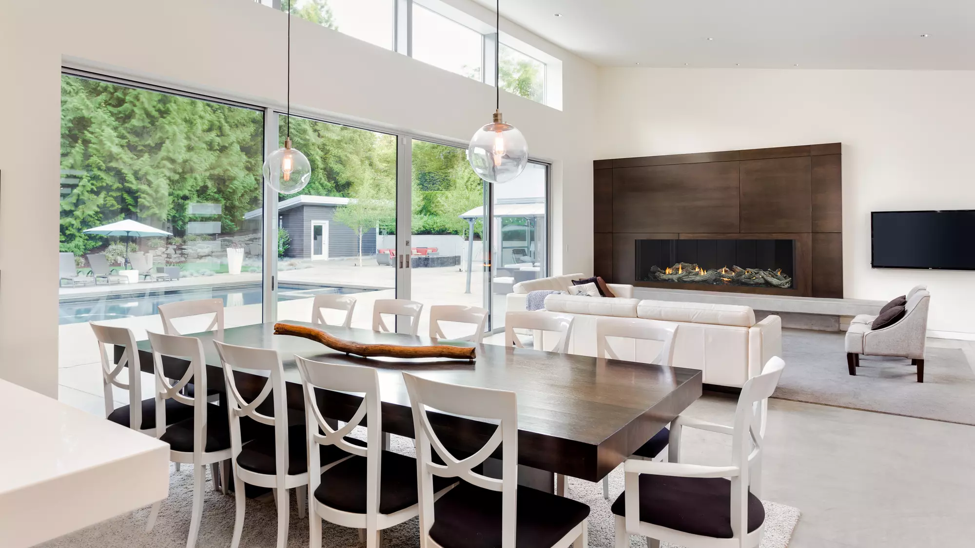 A modern, spacious living room with large sliding glass doors overlooking an outdoor pool includes a sleek dining table, cozy sofas, an Enlight Single-Sided Gas Fireplace by Stellar by Heat & Glo, and a